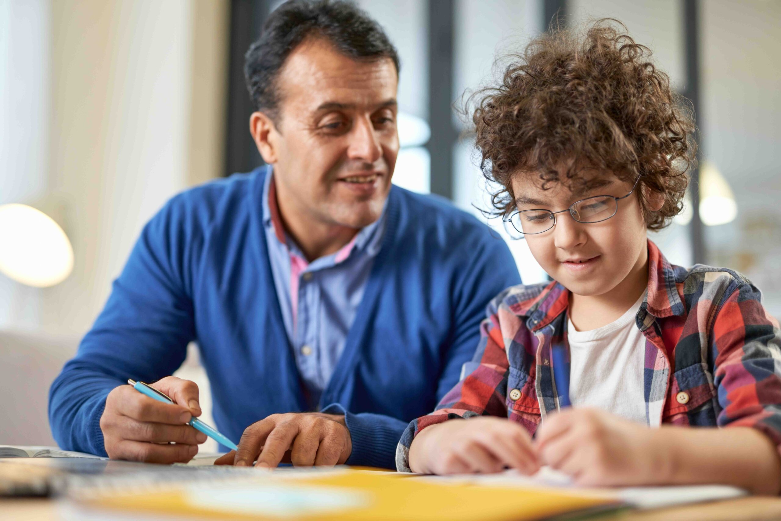 Cute latin boy wearing glasses looking focused while sitting at the desk together with his father and doing homework during remote learning at home. Online education, homeschooling, parenthood